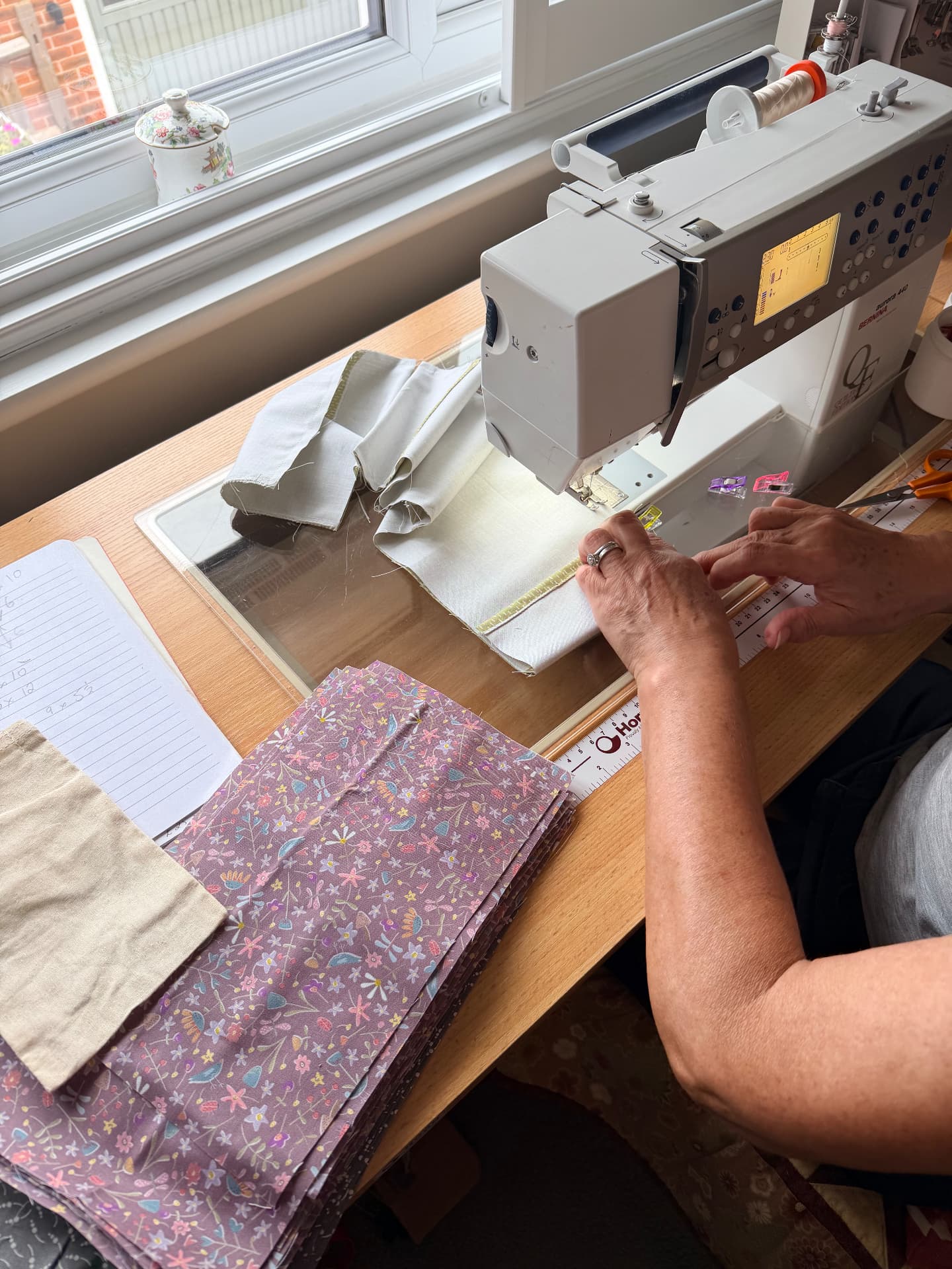 Elaine sewing calico eye pillow covers at a sewing machine by a window, with folded floral fabric and a notebook of measurements beside her.