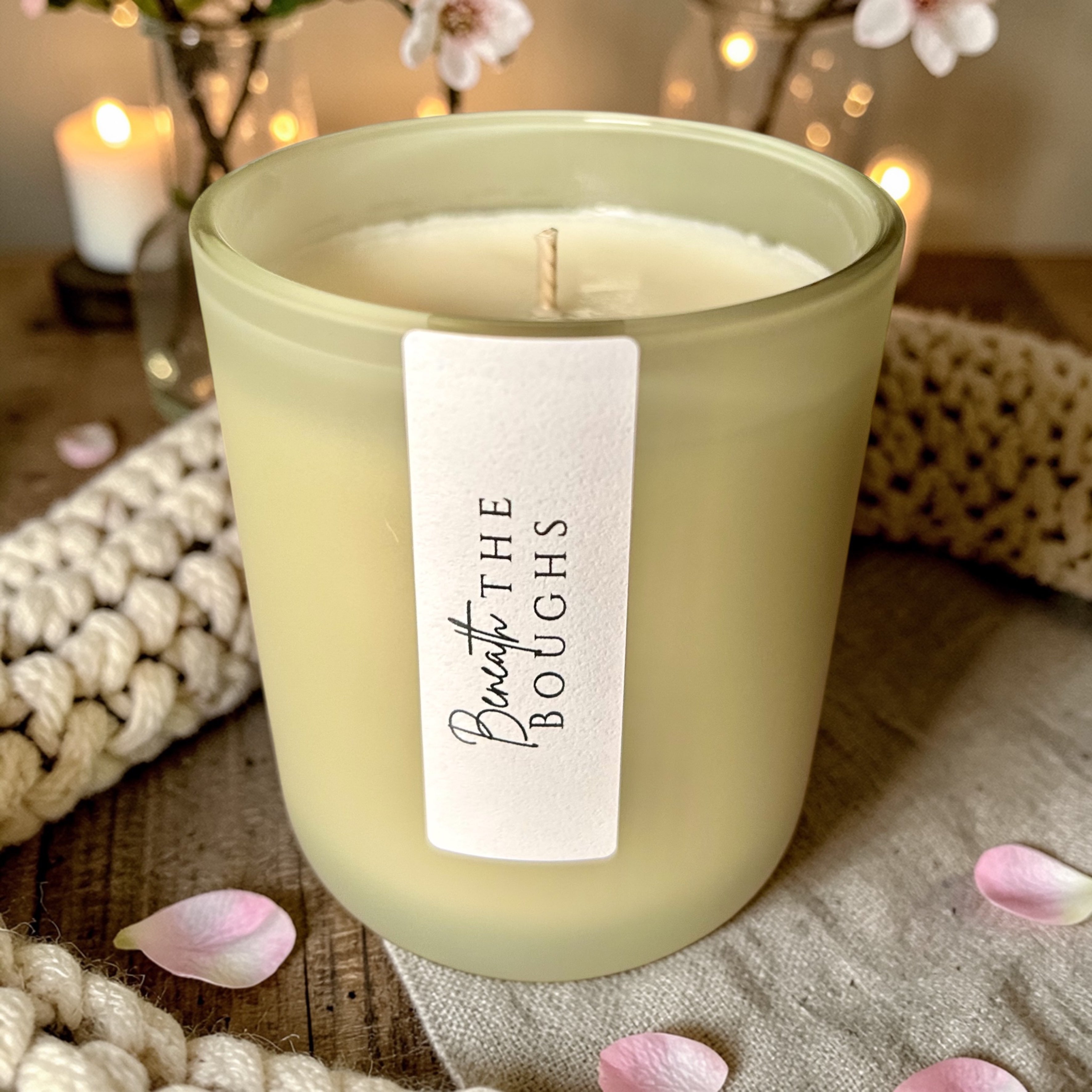 Beneath the Boughs Candle with an unlit wick in a pale green glass jar, surrounded by pink petals and cream knit fabric.