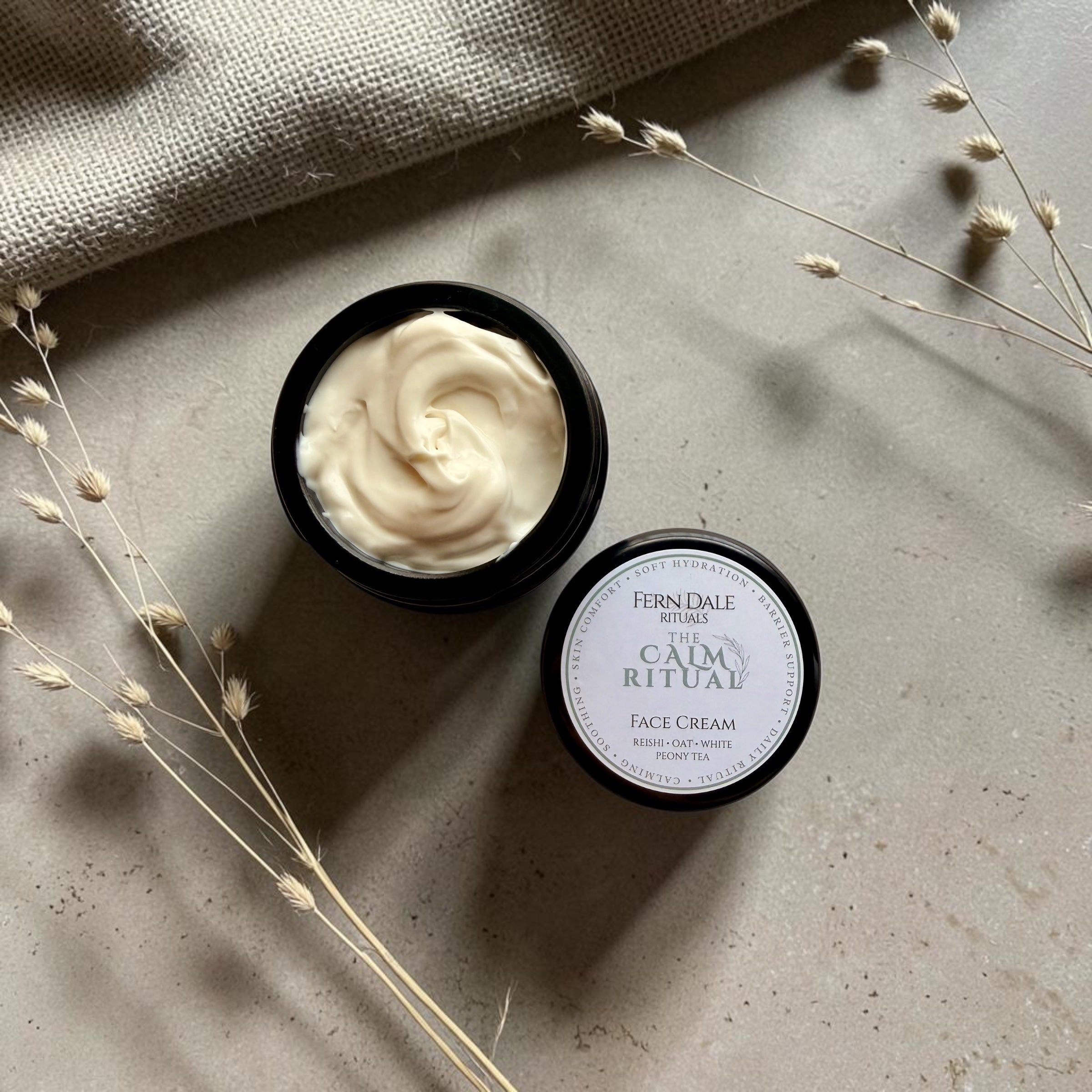 Calm - Face Cream in an open dark jar with pale cream swirls, beside its labelled lid, dried grasses and hessian.