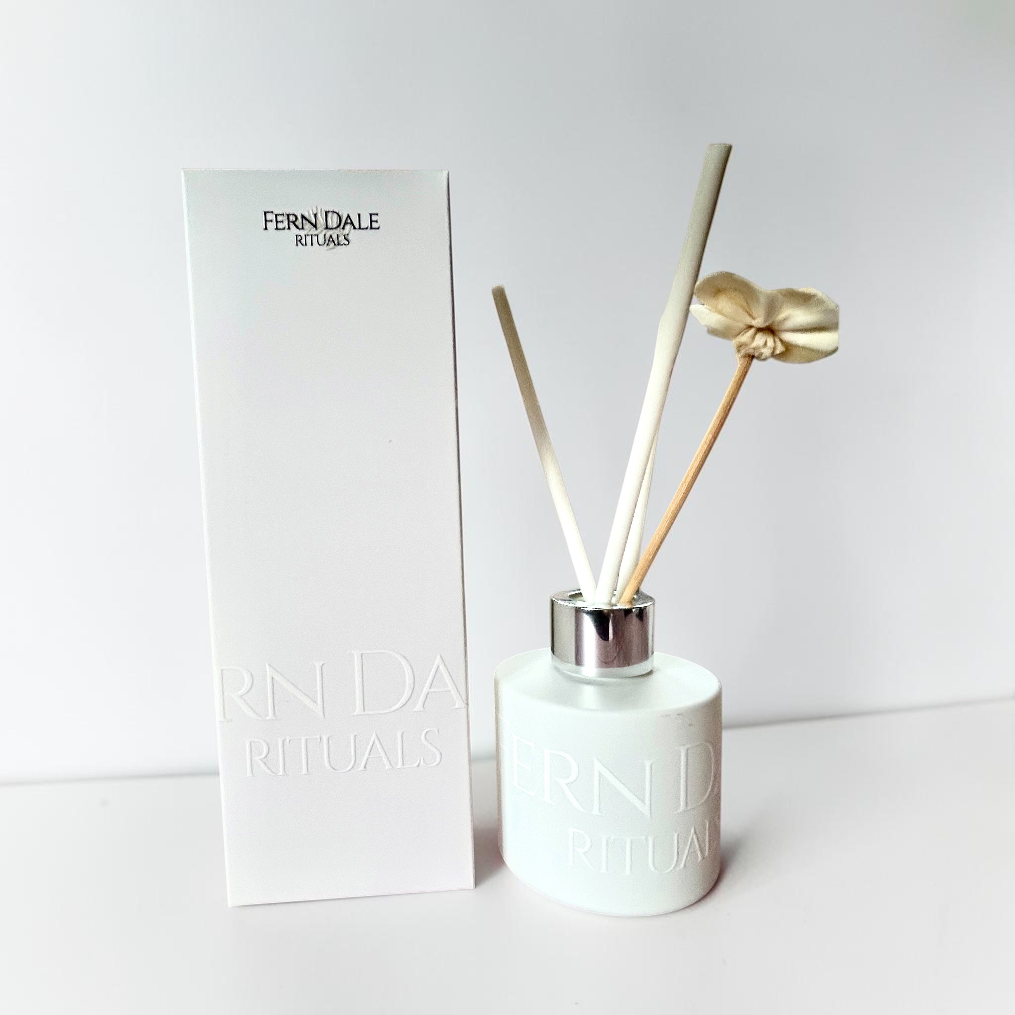 Reed Diffusers white bottle with a silver collar, pale reeds and a cream flower beside a white branded box.