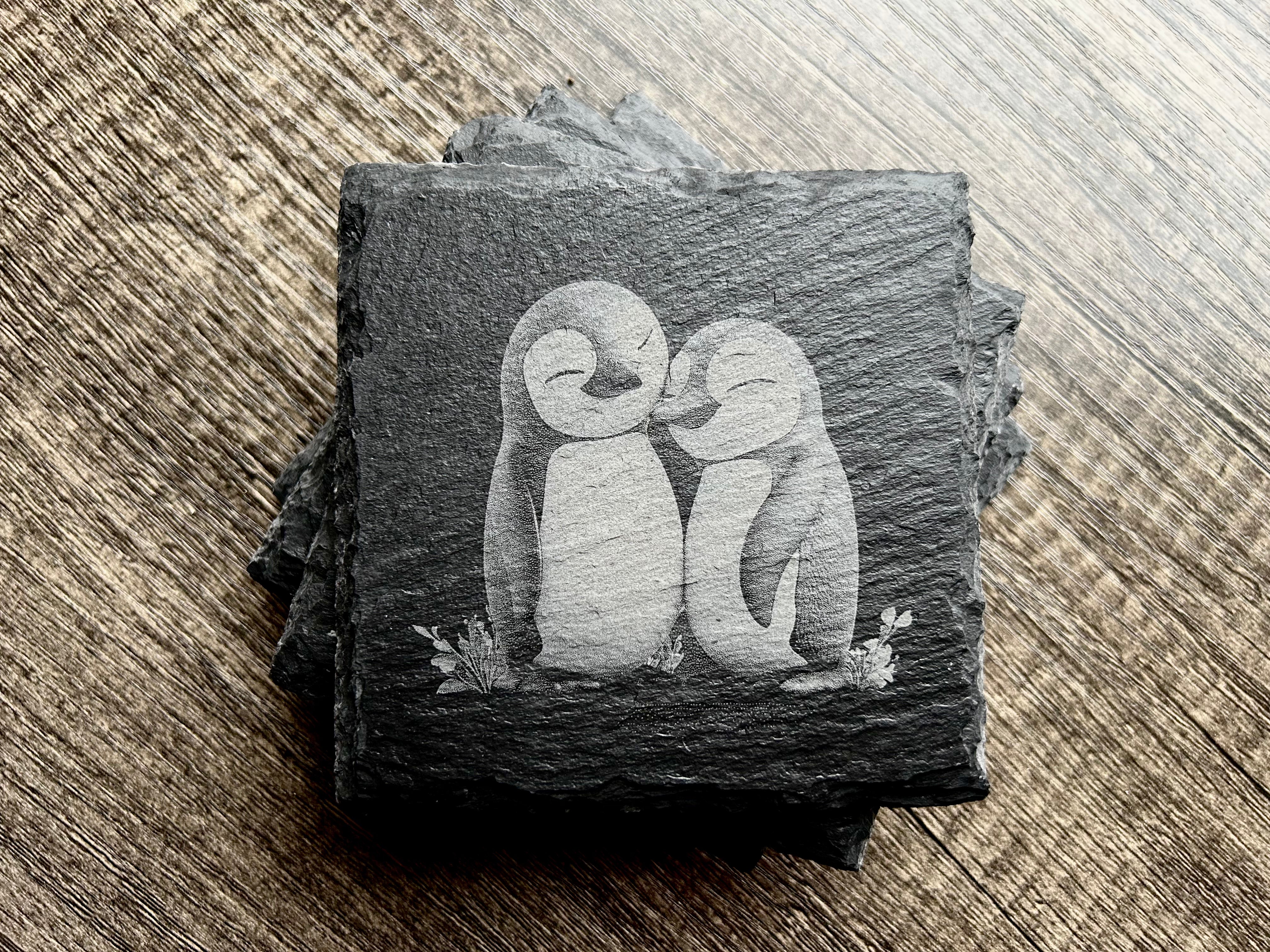 Laser Engraved Slate Coaster Set stacked on wood, with two penguins and small floral sprigs engraved on the top coaster.