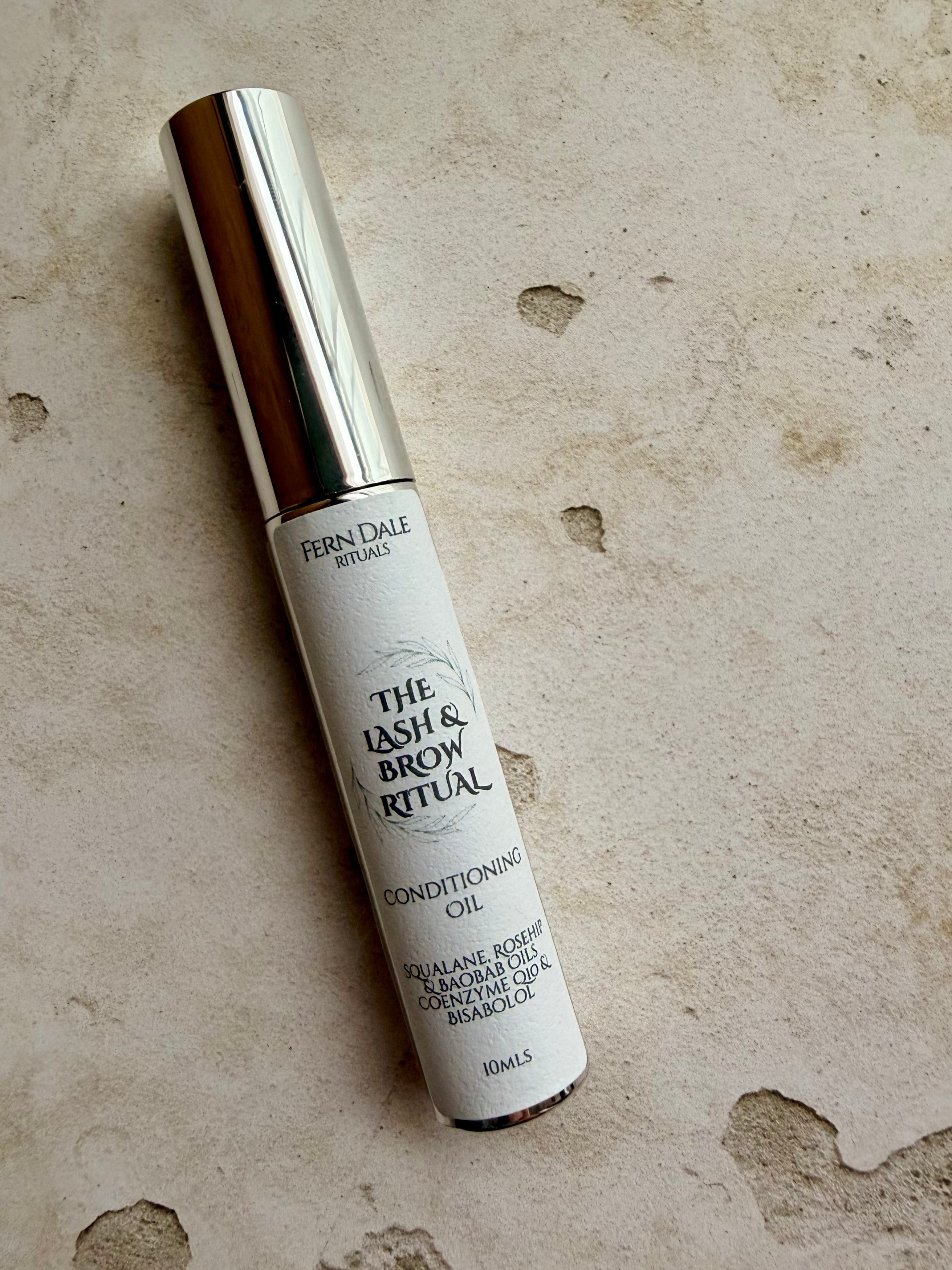 Lash & Brow Conditioning Oil in a slim tube with a silver cap and white label on a textured beige surface.