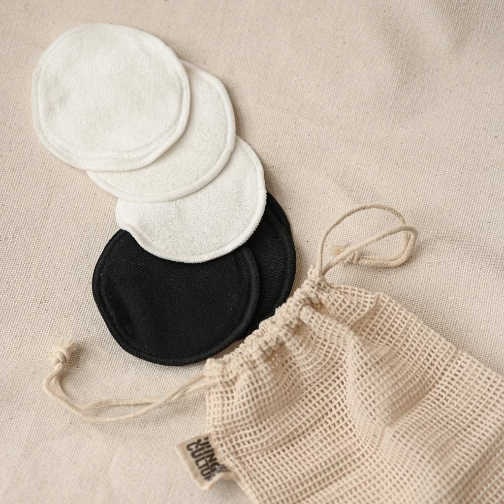Round white and black Make Up Remover Pads beside a beige mesh drawstring bag on cream fabric.