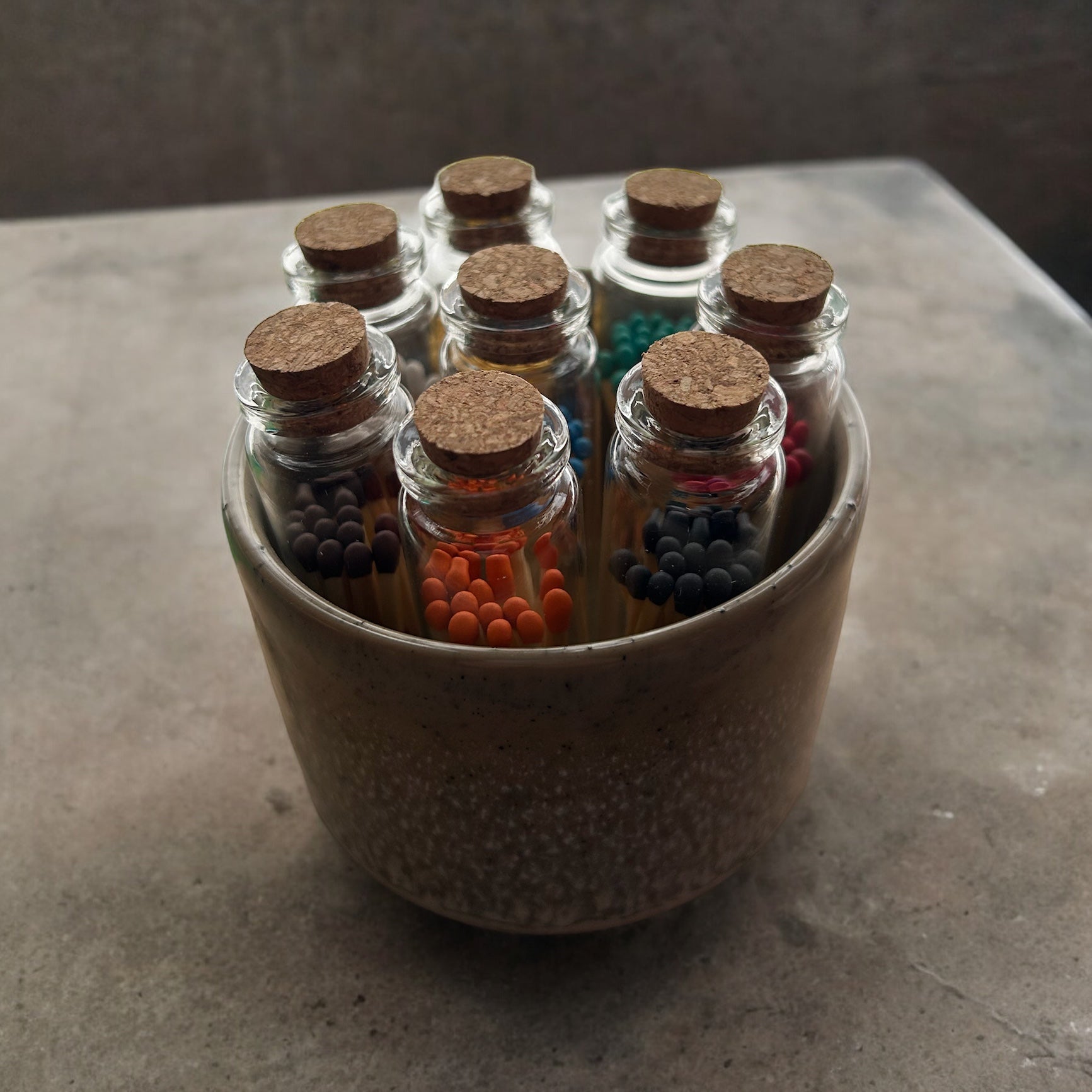 Matches with assorted coloured heads in cork-stoppered glass bottles, arranged in a speckled ceramic pot.