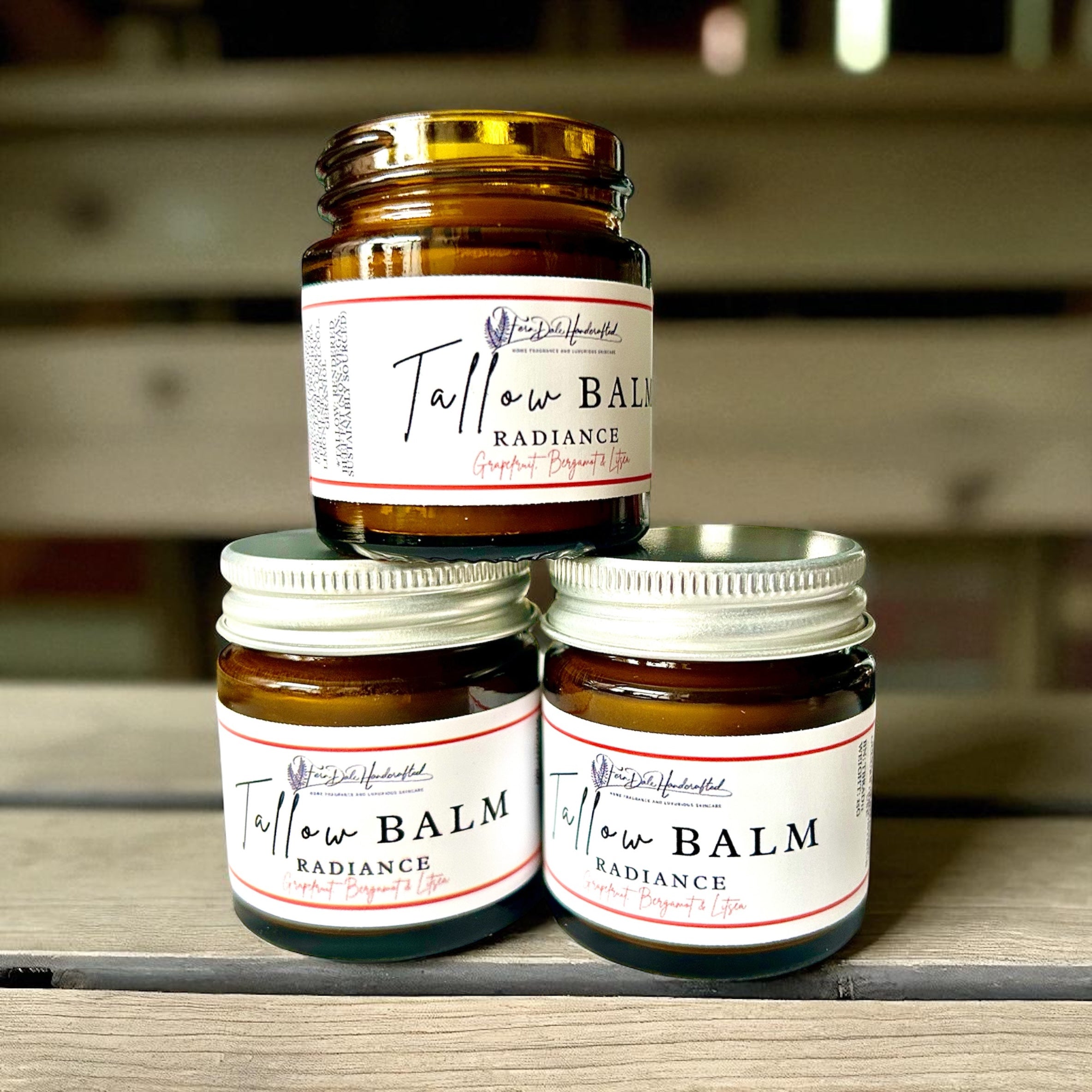Three amber jars of Radiance Tallow Balm stacked on wood, with an open jar above two silver-lidded jars.