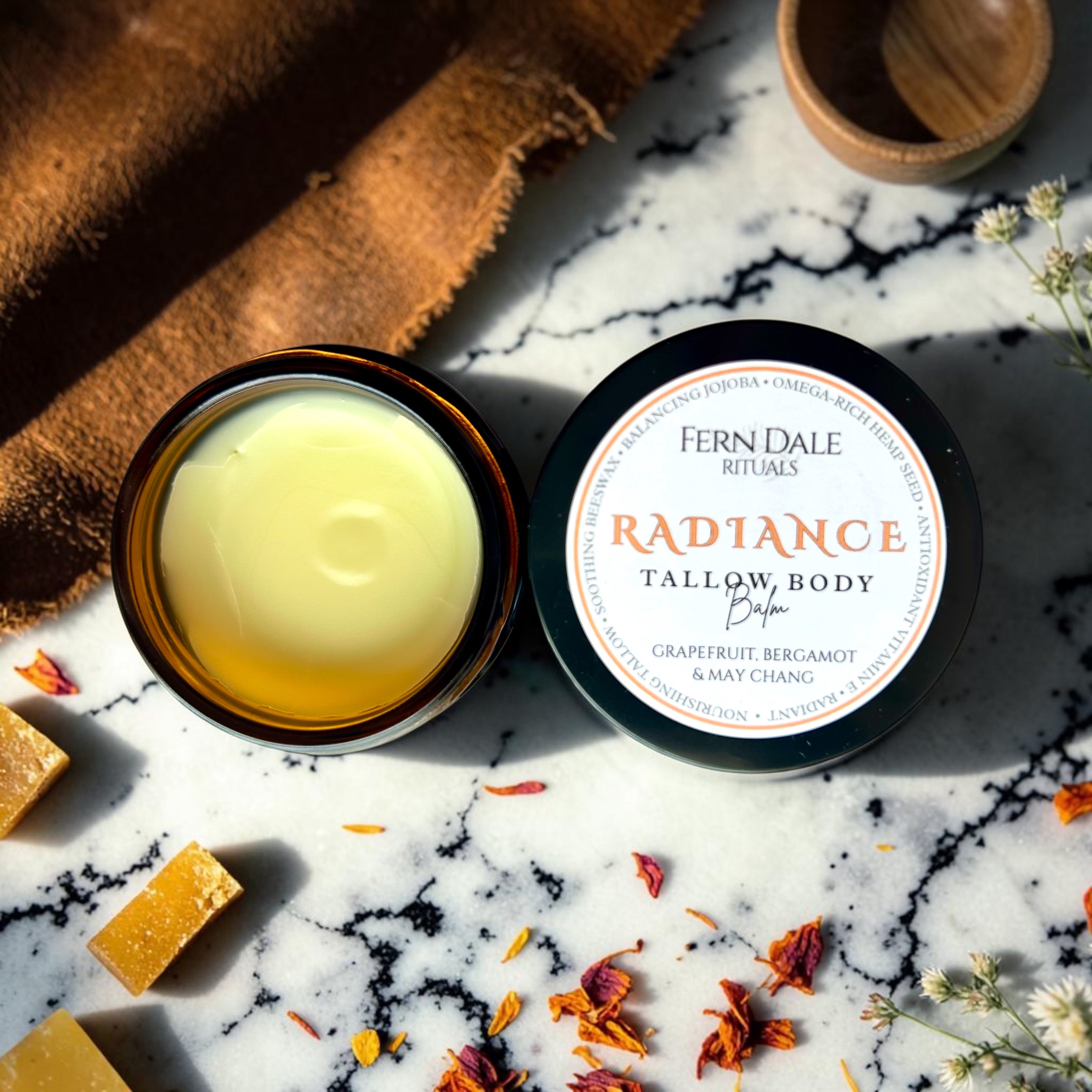 Radiance Tallow Body Balm in an open amber jar beside its black labelled lid on a marble-patterned surface.