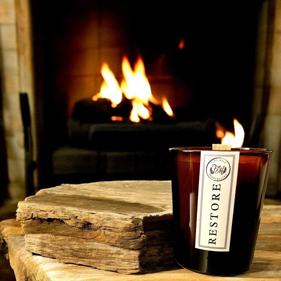Restore candle in an amber glass jar with a white label, beside stacked wood in front of a lit fireplace.