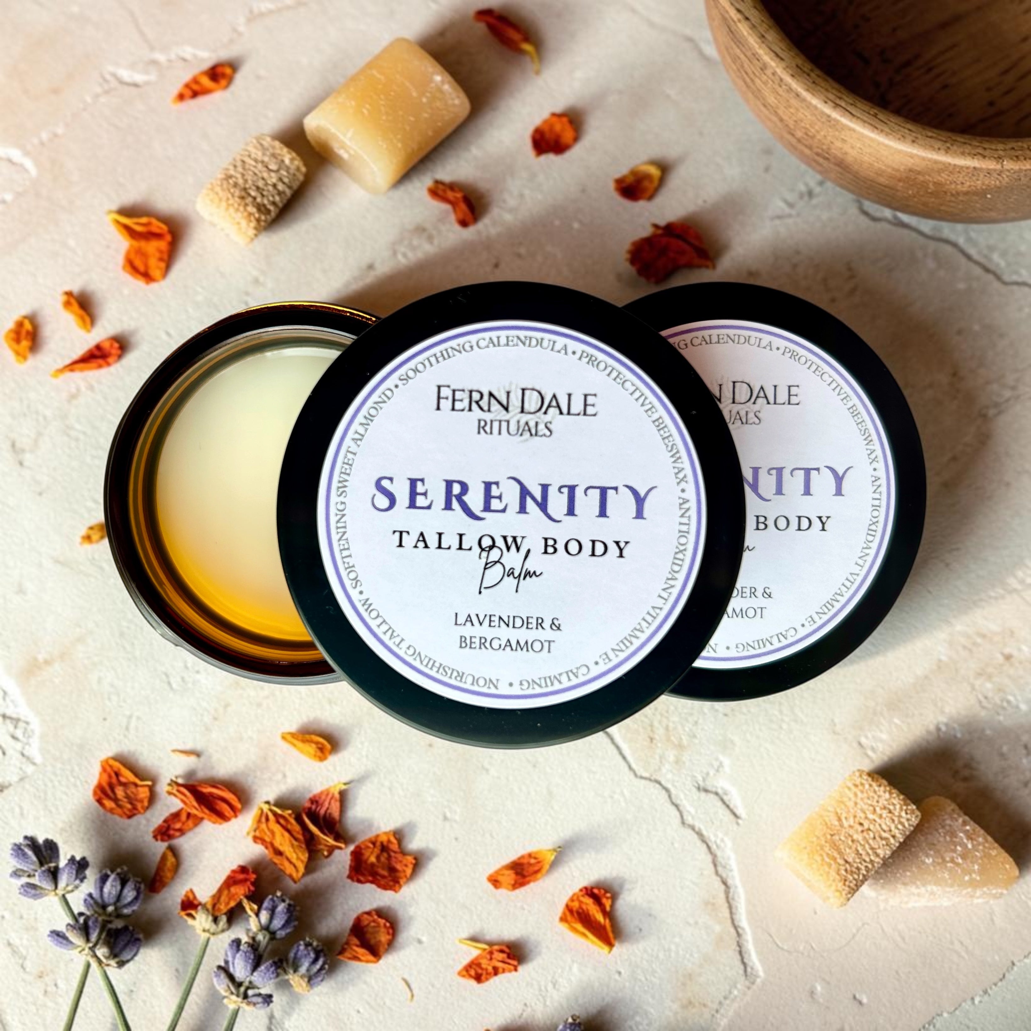 Serenity Tallow Body Balm in an open amber jar with black labelled lids, surrounded by orange petals and purple flowers.