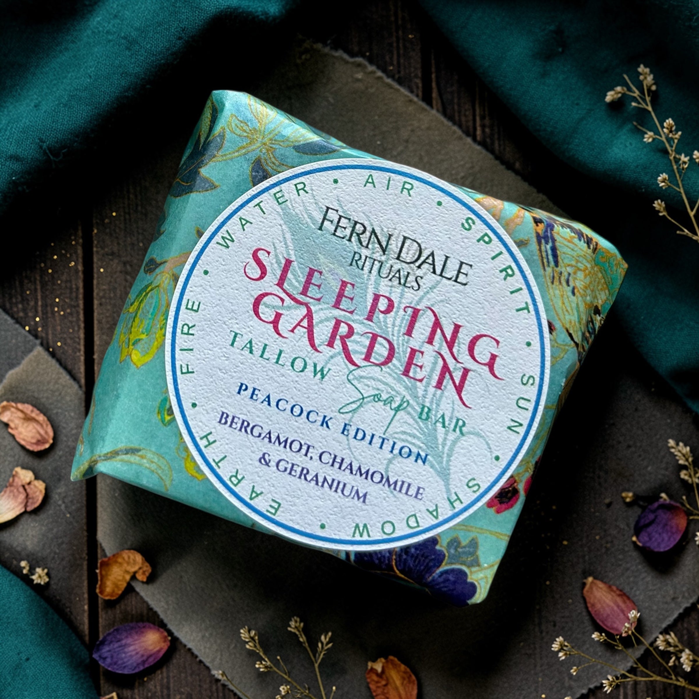 Sleeping Garden Tallow Soap - Peacock Edition in turquoise patterned wrapping with a round white label, amid dried petals.