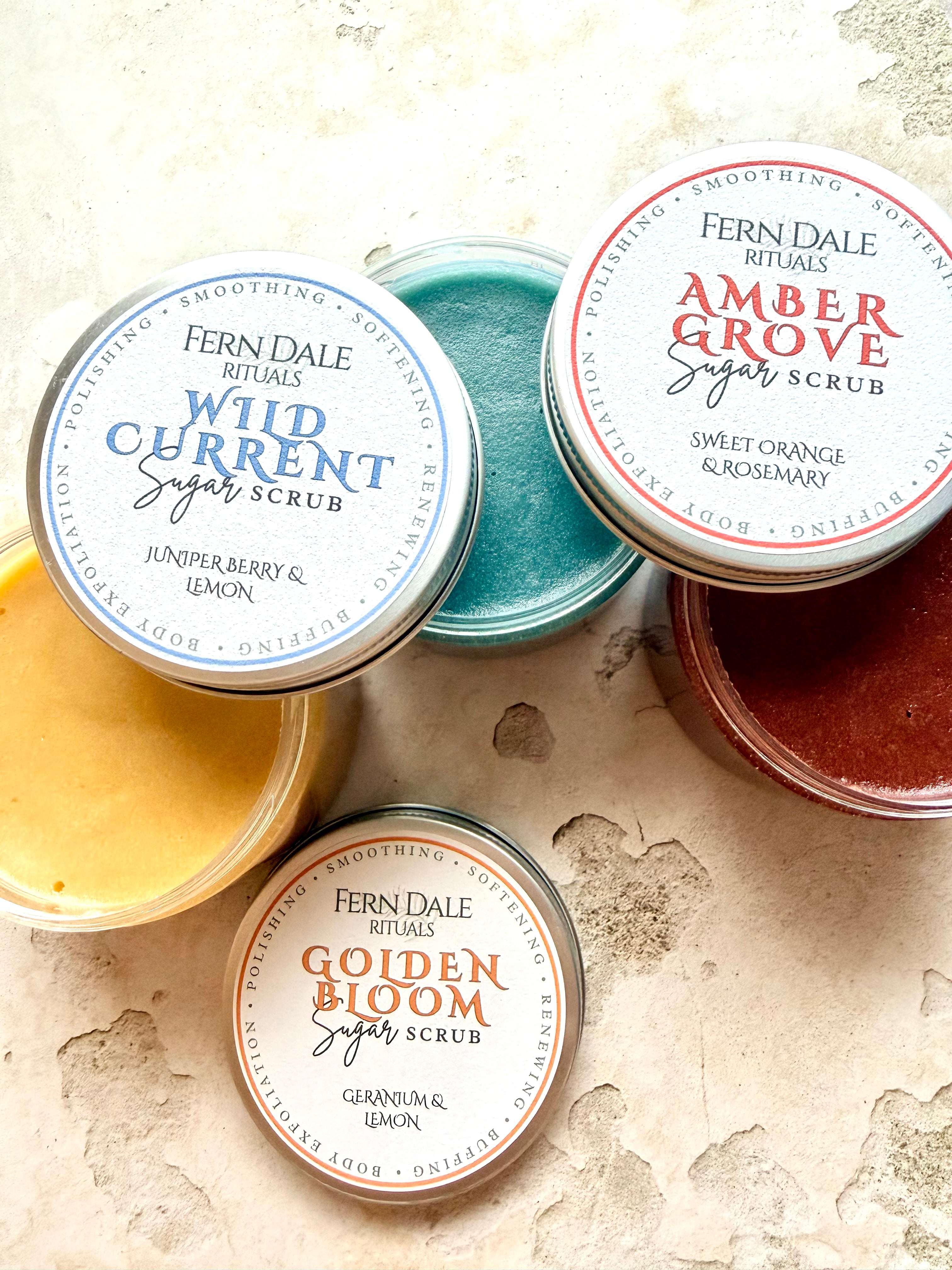 Sugar Scrubs in open clear jars showing yellow, turquoise and red textures, with labelled silver lids.