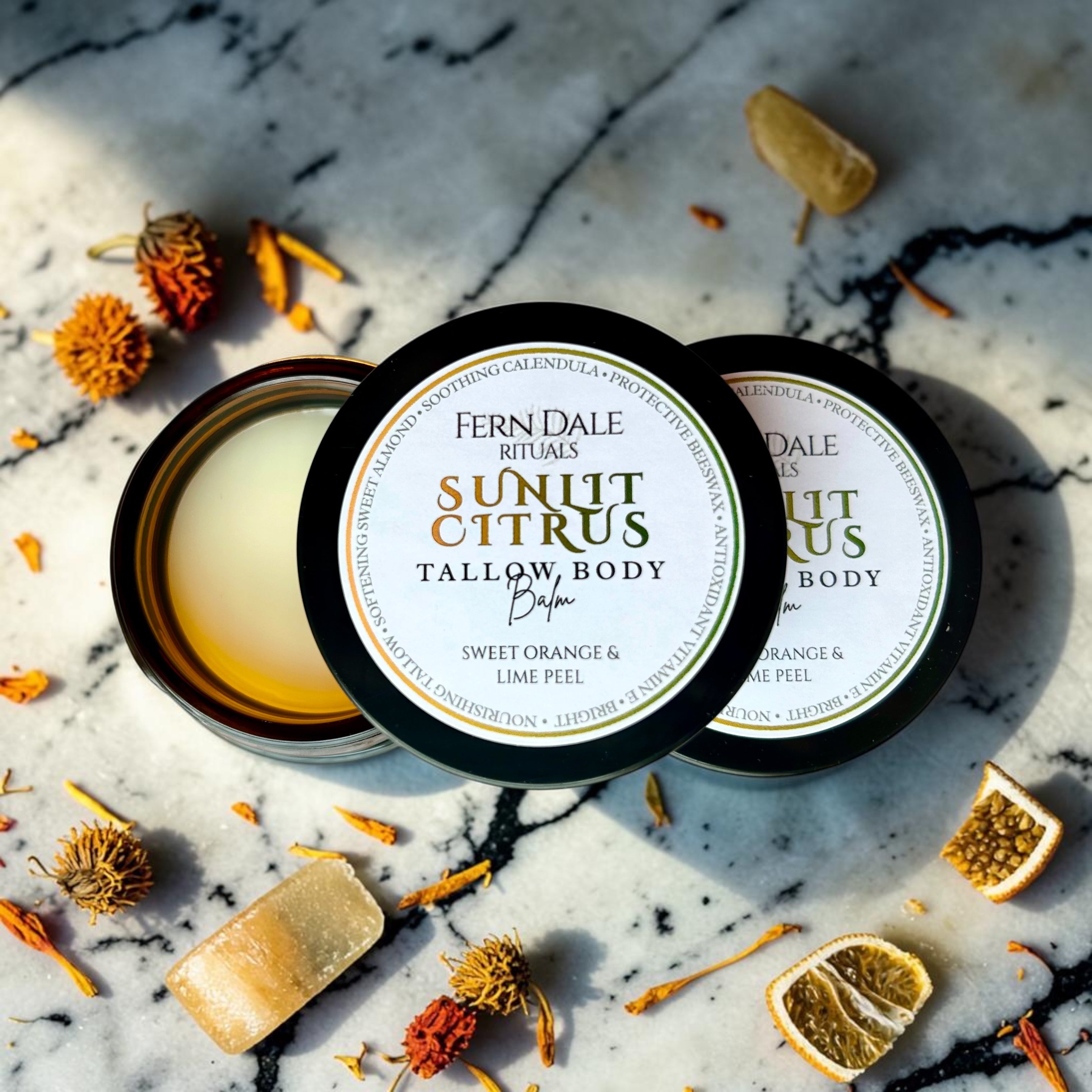Sunlit Citrus Tallow Body Balm in open and closed jars with black lids, surrounded by dried flowers and citrus pieces.