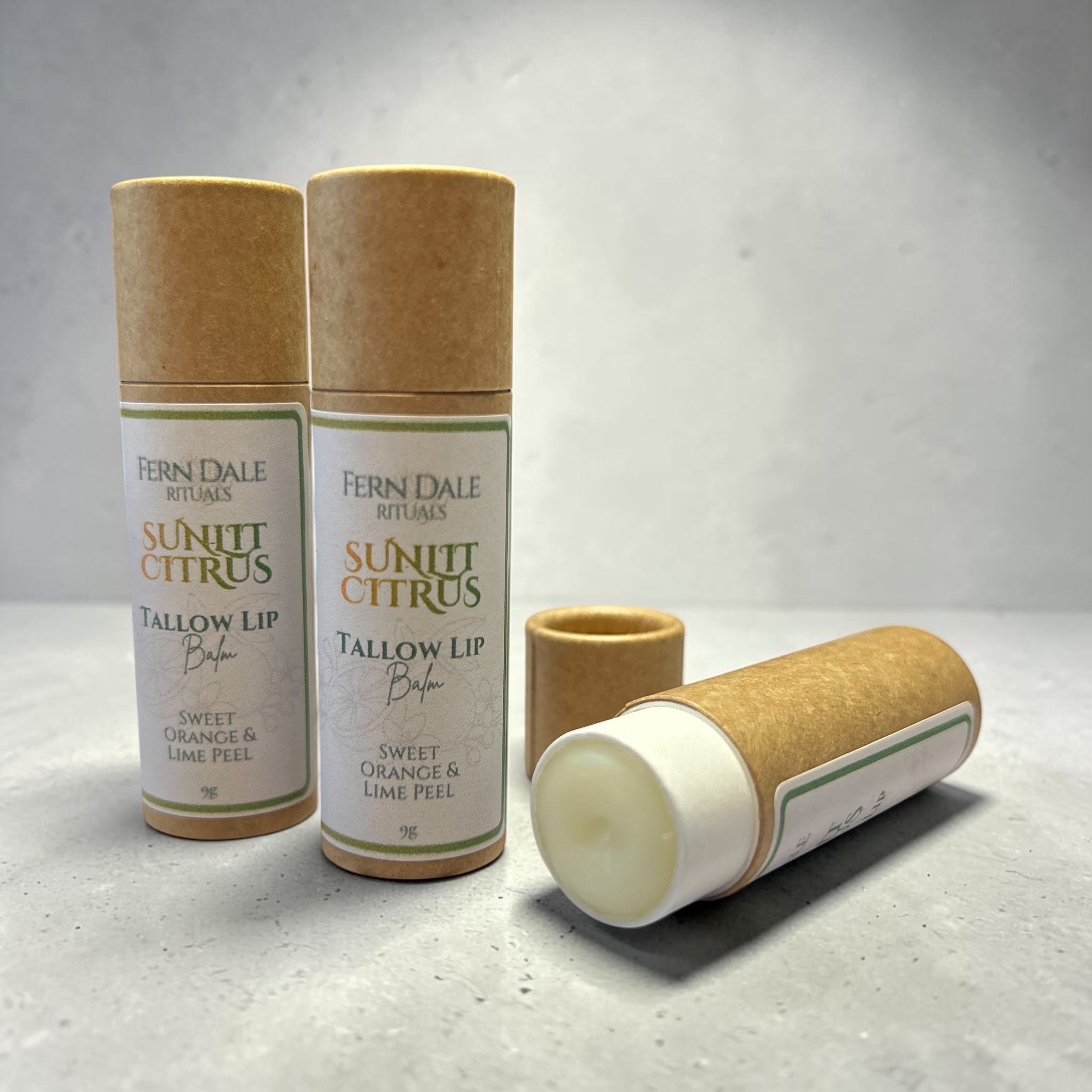 Sunlit Citrus Tallow Lip Balm in three brown cardboard tubes, two standing capped and one lying open with pale balm visible.
