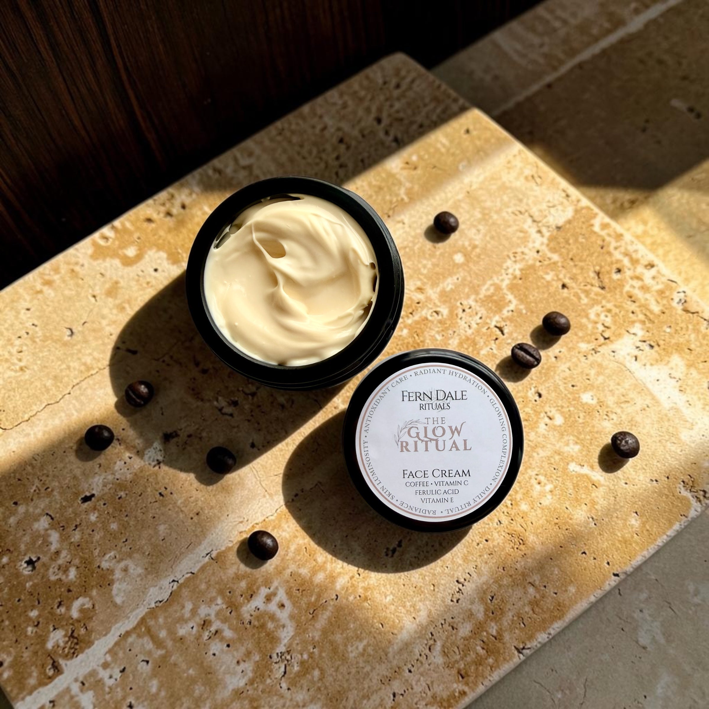 The Glow Ritual - Face Cream in an open black jar beside its labelled lid and coffee beans on sunlit stone.