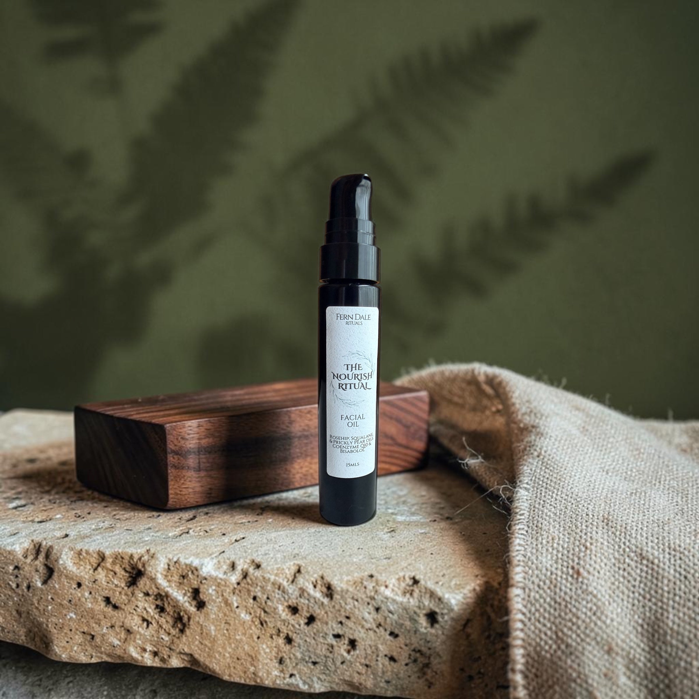 The Nourish Ritual - Facial Oil in a black pump bottle with a white label, beside a wooden block and beige fabric.