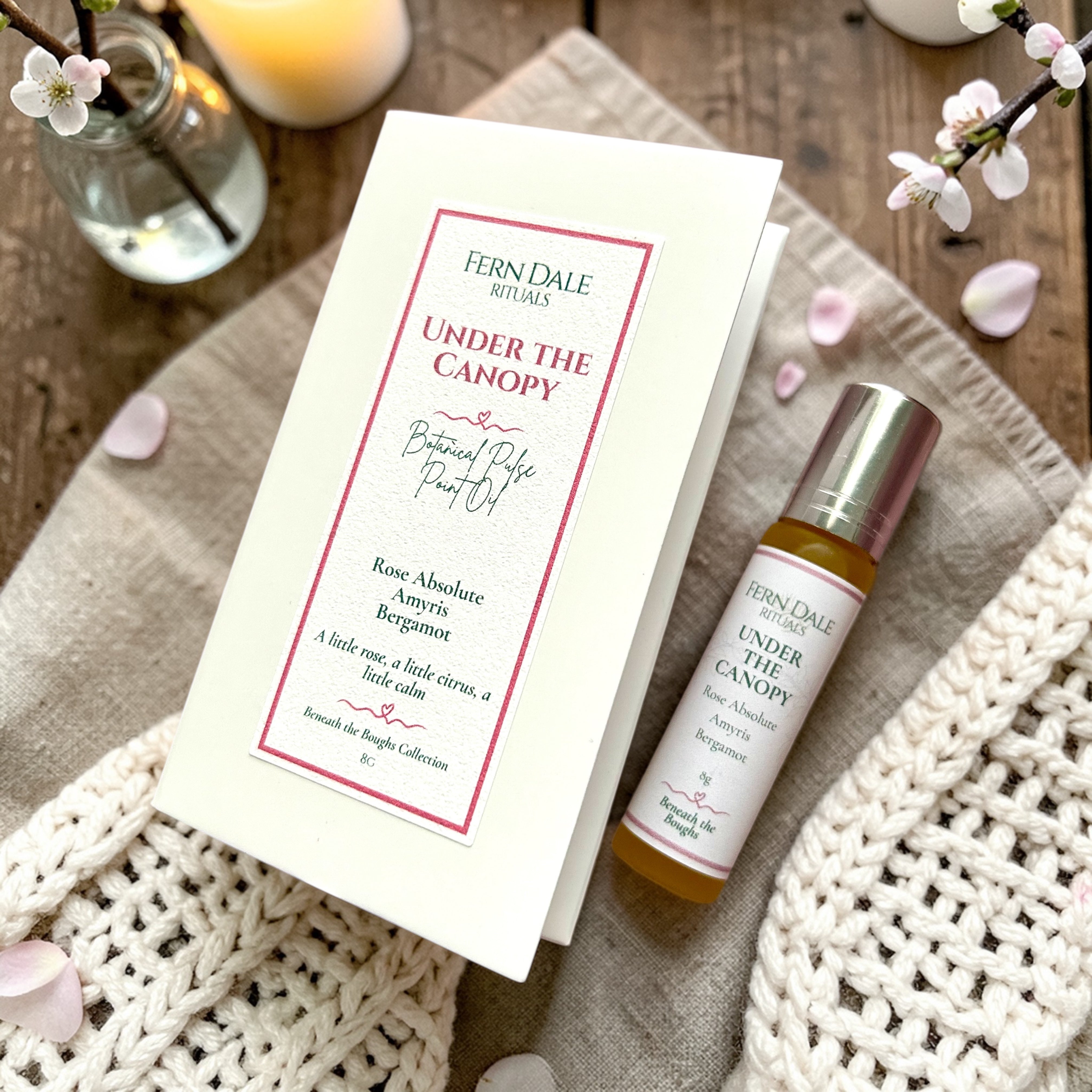 Under the Canopy - Botanical Rose Pulse Point Oil bottle with a silver cap beside cream packaging and pink petals.