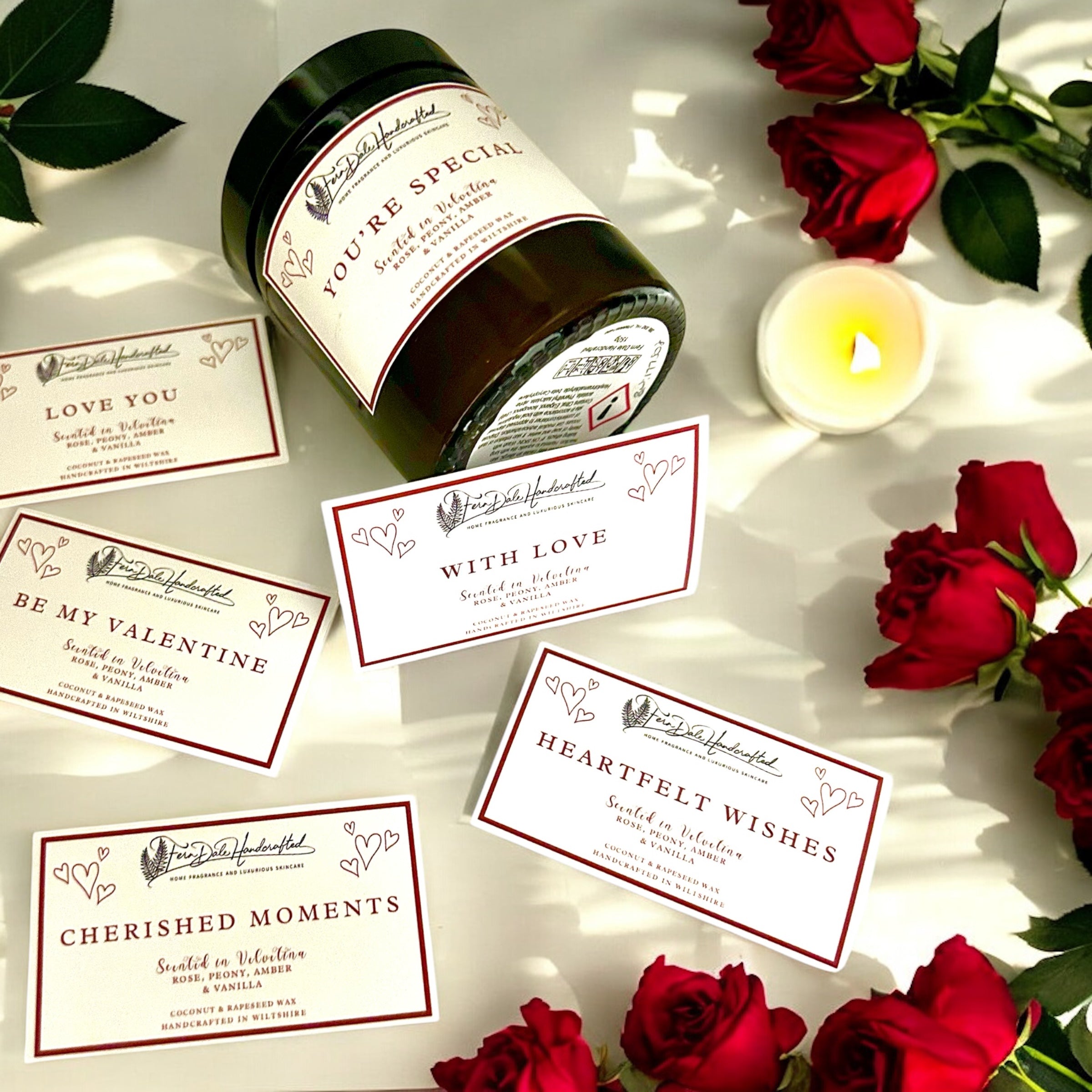 Velvetina Candle in a dark jar with a black lid, surrounded by red roses, message labels and a lit tealight.