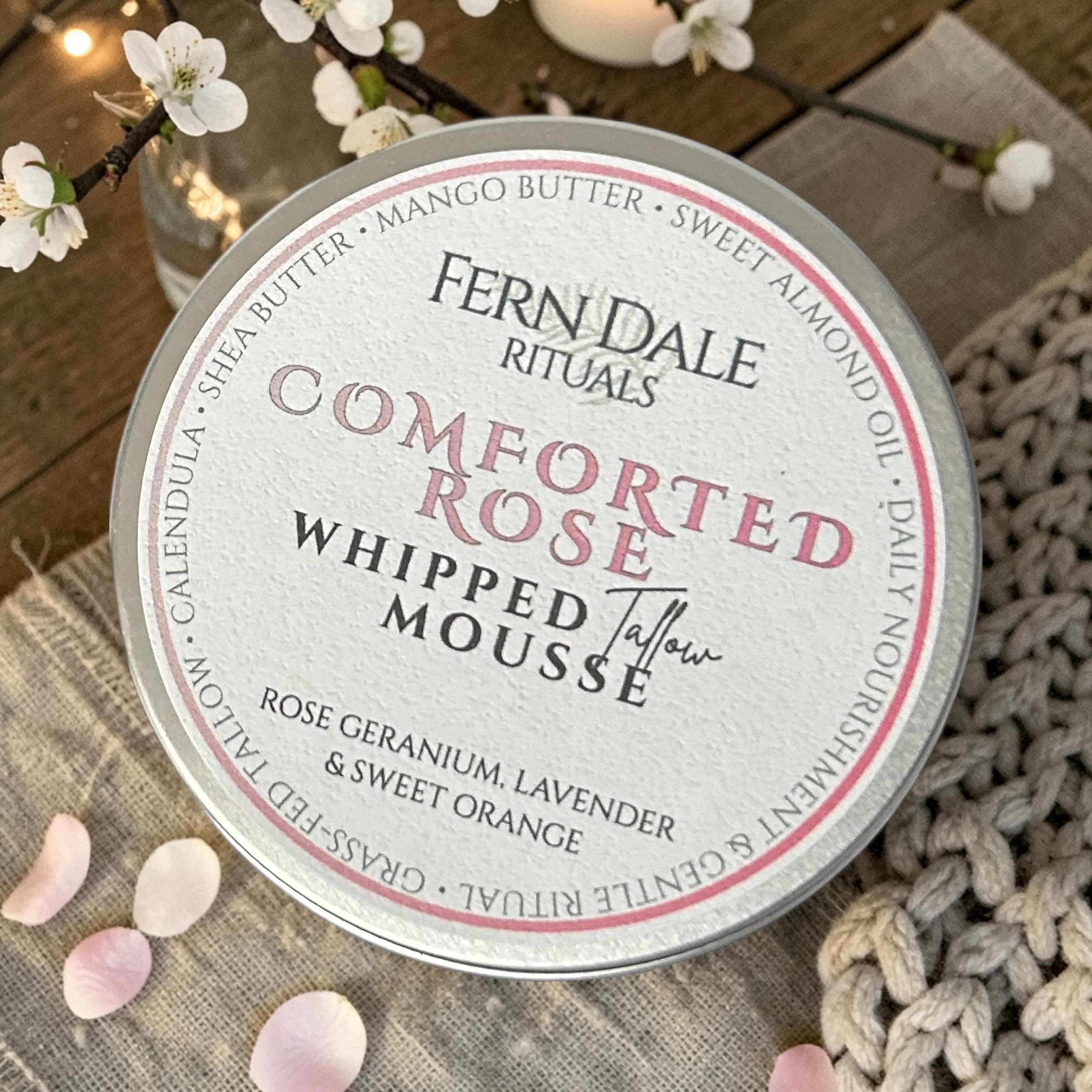Whipped Tallow Mousse - Comforted Rose tin with a pink-bordered label, beside pale pink petals and white blossom.